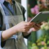 Florist in apron using digital tablet inside flower shop with blurred floral background.