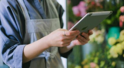 Florist in apron using digital tablet inside flower shop with blurred floral background.