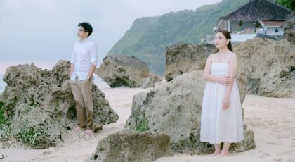 Young couple stands amongst rocks on tropical beach, contemplating view.