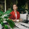 Woman sipping coffee while working on a laptop indoors with lush greenery.