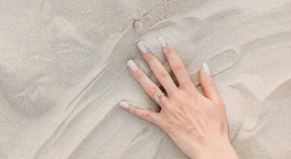 A close-up of a female hand with a diamond ring and manicured nails on sand.