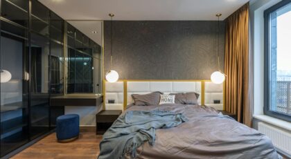 Modern light bedroom interior with soft bed with pillows and blanket near lamps and mirror with shelf near pouf and window with curtains