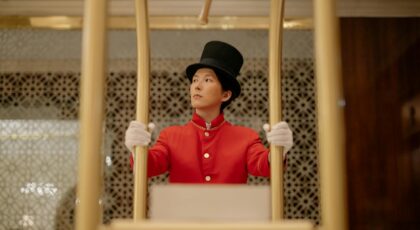 A bellboy in a red uniform stands with a luggage cart inside a luxurious hotel.